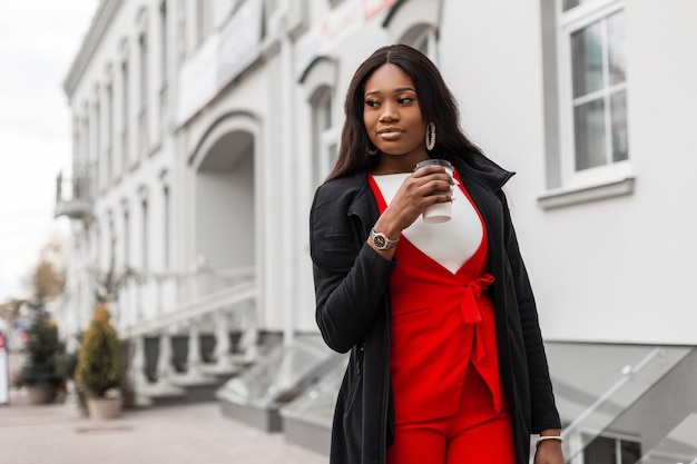 Mode-Modell ziemlich feine junge afrikanische Frau im stilvollen roten Anzug in Jacke mit Tasse Kaffee Spaziergänge in der Nähe von Vintage-Gebäude in der Stadt. Modisches sexy schwarzes Mädchen genießt leckeres Getränk und geht im Freien.