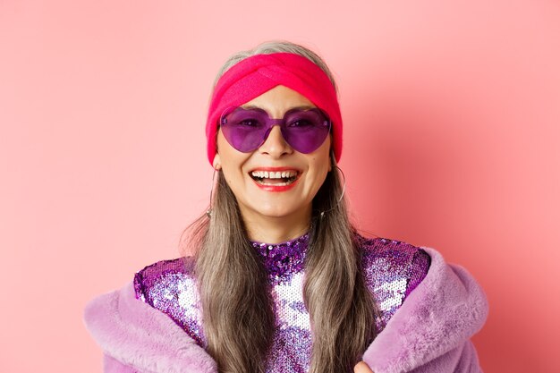 Mode. Nahaufnahme einer stilvollen Seniorin, die glücklich in die Kamera lächelt, Sonnenbrille und Disco-Stirnband trägt, rosa Hintergrund.