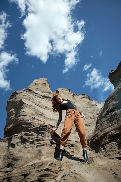 Mode rothaarige Frau in orange Jeans, die in der Natur nahe sandigen Felsen aufwirft