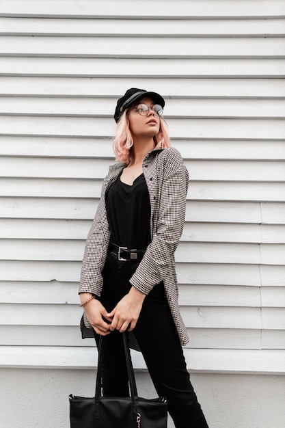 Mode stilvolle schöne junge Frau mit Brille und Hut in trendiger Freizeitkleidung mit einer Modetasche steht in der Nähe einer Holzwand