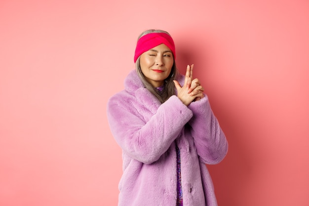 Mode- und Einkaufskonzept. Coole und stilvolle asiatische Seniorin in lila Kunstpelzmantel, die eine Fingergewehrgeste macht und mit einem frechen Lächeln nach links schaut und sich wie ein Geheimagent verhält, rosa Hintergrund.