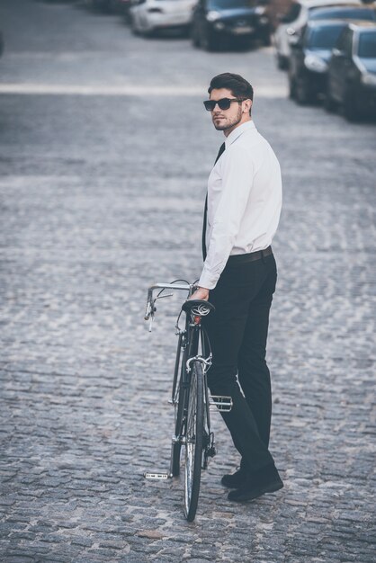Mode unterwegs. Rückansicht in voller Länge eines gutaussehenden, gut gekleideten jungen Mannes mit Sonnenbrille, der sein Fahrrad im Freien rollt
