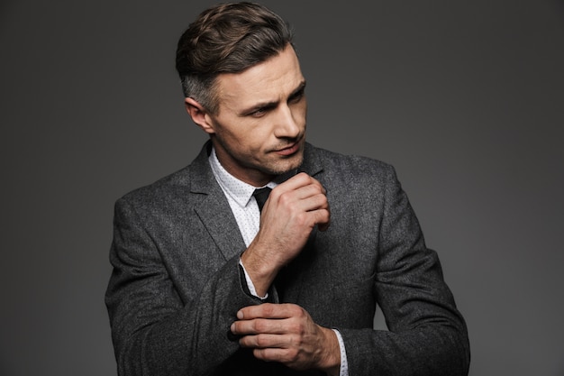 Modebild des maskulinen Mannes gekleidet im Geschäftsanzug, der beiseite schaut, während Manschettenknopf oder Knopf auf Ärmel der Jacke, lokalisiert über graue Wand befestigt