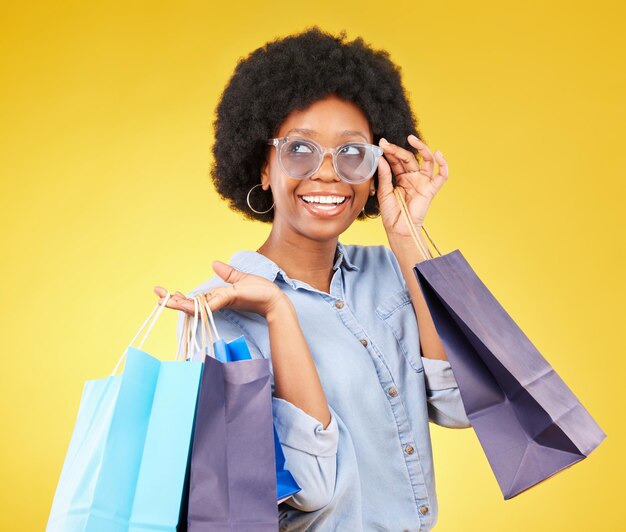 Modedenkende und schwarze Frau mit Einkaufstüten lächeln und freuen sich auf den Verkauf im Studio auf gelbem Hintergrund. Shopper-Mädchen und coole Kundin sind fröhlich für Boutique-Einzelhandel oder Ladenrabatt