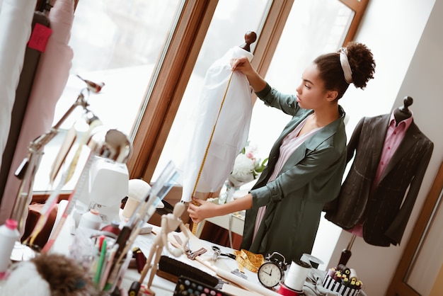 Modedesigner bei der Arbeit. Junge afrikanische Frau, Schneiderdesignerin schaut auf die Schaufensterpuppe mit T-Shitrt. Sie misst Kleidung