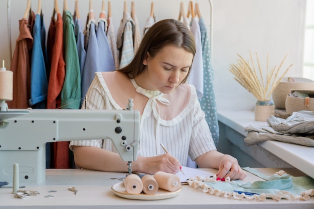 Modedesignerin, die Kleidungsskizze zeichnet, während sie an ihrem Arbeitsplatz mit Nähmaschine sitzt