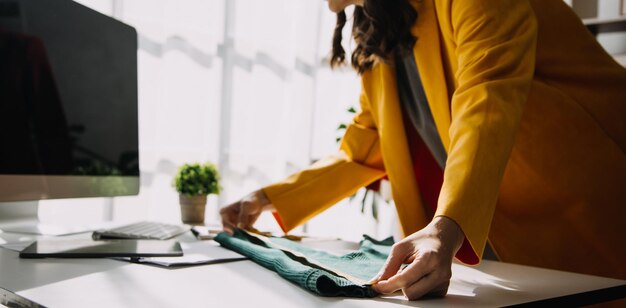 Modedesignerin l Junge asiatische Frau, die mit einem Laptop-Tablet arbeitet und lächelt, während sie in der Werkstatt steht und geschäftlich reagiert