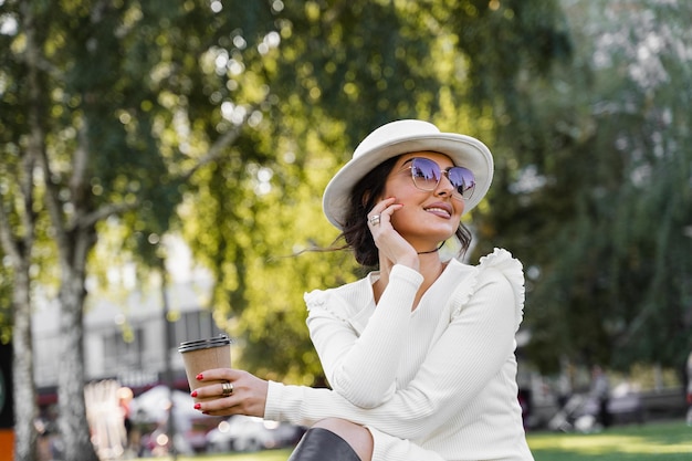 Modefrau genießt Kaffee. Attraktives Model trug weißes Kleid, Hut und Sonnenbrille und posierte auf der Straße.