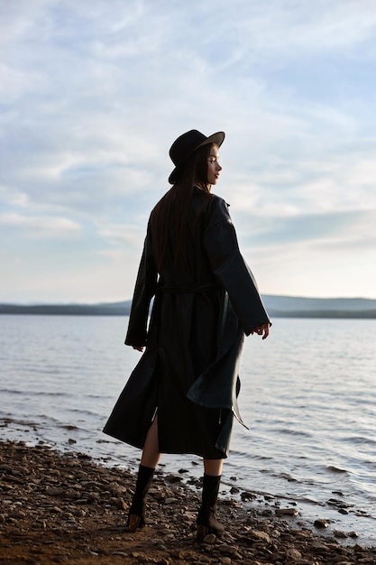 Modefrau im schwarzen runden Hut und im ledernen Regenmantel nahe Strandseewinter. Schönes Make-up und leuchtend roter Lippenstift auf den Lippen