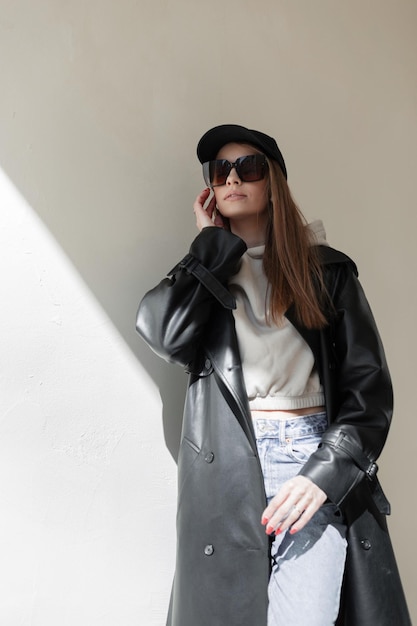 Modehübscher Frauen-Hipster mit Sonnenbrille und schwarzer Mock-up-Mütze in schwarzem Ledermantel mit Goodie und Jeans steht in der Nähe einer Wand auf der Straße