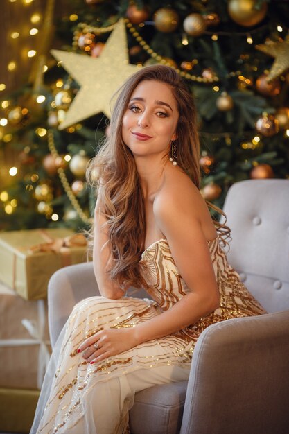 Modeinnenfoto der schönen sinnlichen Frau mit dem blonden Haar im luxuriösen goldenen Kleid, das neben einem Weihnachtsbaum aufwirft. Wünschen Ihnen ein frohes Weihnachtsfest.