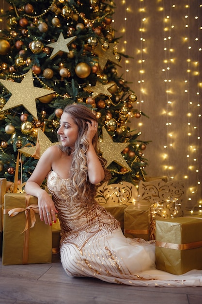 Modeinnenfoto der schönen sinnlichen Frau mit dem blonden Haar im luxuriösen goldenen Kleid, das neben einem Weihnachtsbaum aufwirft. Wünschen Ihnen ein frohes Weihnachtsfest.