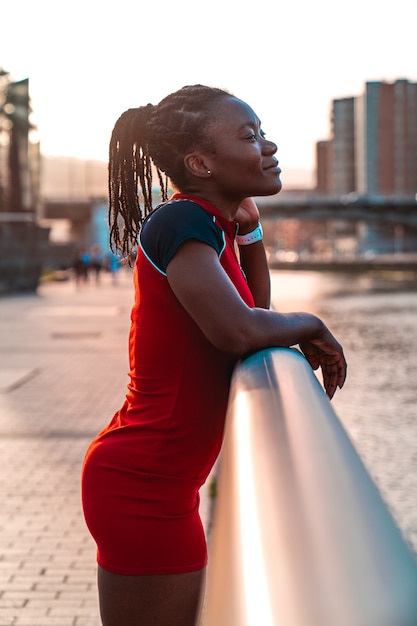 Model Mädchen mit schwarzer Haut und Afro-Stil Haaren posiert lächelnd am Geländer einer Brücke am Fluss einer Stadt in einem wunderschönen Sonnenuntergang mit warmen Farben in einem roten Kleid festhalten