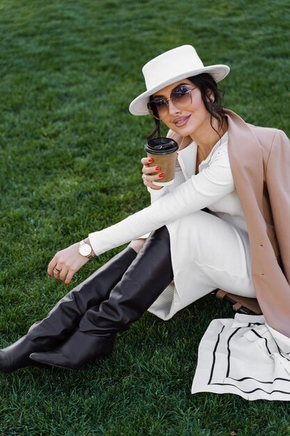 Model mit Kaffeetasse auf dem Rasen. Attraktive Frau mit Hut, Kleid, Jacke und Sonnenbrille.