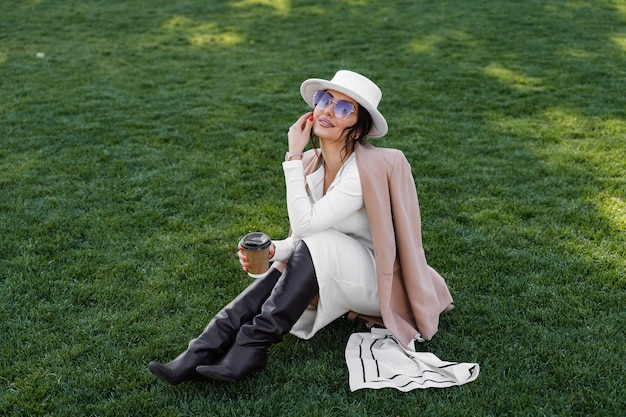 Model mit Kaffeetasse auf dem Rasen. Attraktive Frau mit Hut, Kleid, Jacke und Sonnenbrille.