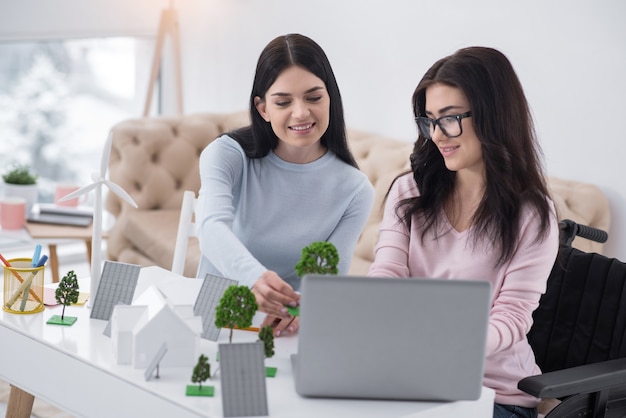 Modelado detallado. Mujer inmóvil optimista y colega usando laptop mientras mira el modelo de árbol