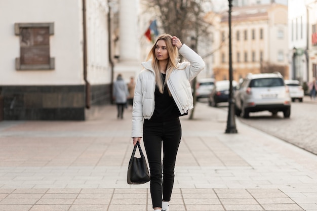 Modell einer jungen Frau in einer weißen Jacke und einer trendigen Ledertasche steht auf der Straße
