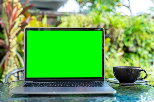 Modell eines Laptop-Computers mit leerem Bildschirm mit Kaffeetasse und Smartphone auf dem Tisch des Cafés im Freien HintergrundGrüner Bildschirm