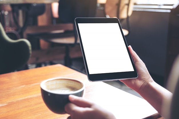 Modellbild der Frauenhände, die schwarzen Tablett-PC mit leerem weißen Bildschirm mit Kaffeetasse auf Holztisch im Café halten