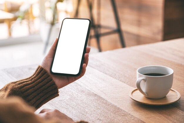 Modellbild der Frauenhand, die ein schwarzes Handy mit leerem Bildschirm mit Kaffeetasse auf Holztisch hält