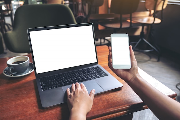 Modellbild der Geschäftsfrau, die Handy mit leerem weißen Bildschirm hält, während Laptop auf Holztisch im Café verwendet