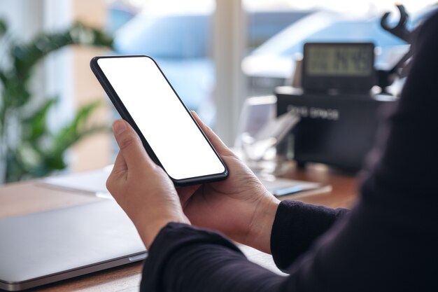 Modellbild der Hände, die schwarzes Mobiltelefon mit leerem weißen Bildschirm mit Laptop auf Holztisch im Büro halten