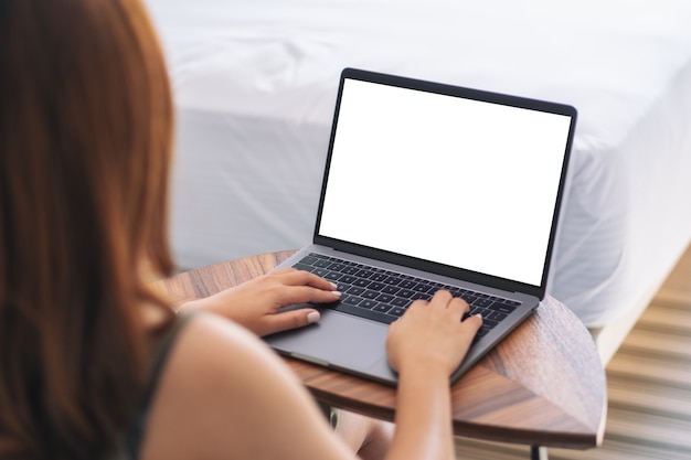 Modellbild einer Frau, die auf Laptop mit leerer weißer Desktop-Bildschirmtastatur auf Holztisch neben dem Bett verwendet und tippt