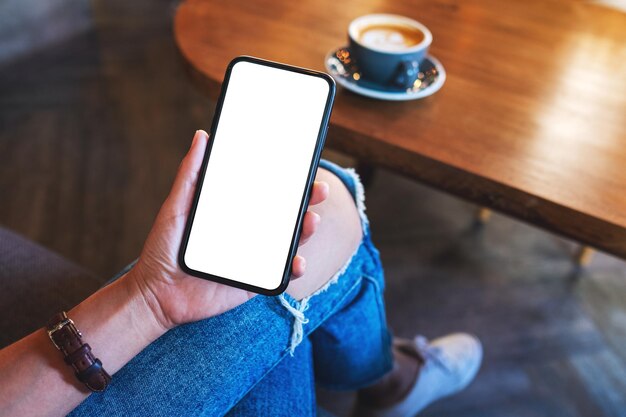 Modellbild einer Frau, die ein Mobiltelefon mit einem leeren weißen Desktop-Bildschirm mit einer Kaffeetasse auf dem Tisch hält