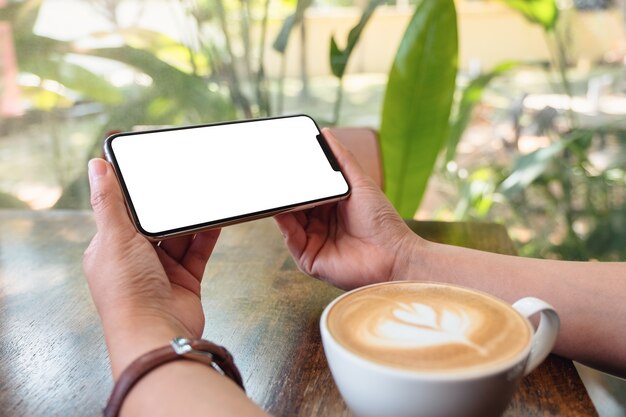 Modellbild einer Frau, die ein schwarzes Handy mit leerem Desktop-Bildschirm mit Kaffeetasse auf Holztisch hält und benutzt