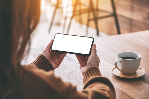 Modellbild einer Frau, die ein schwarzes Mobiltelefon mit einem leeren Desktop-Bildschirm horizontal mit einer Kaffeetasse auf dem Tisch hält