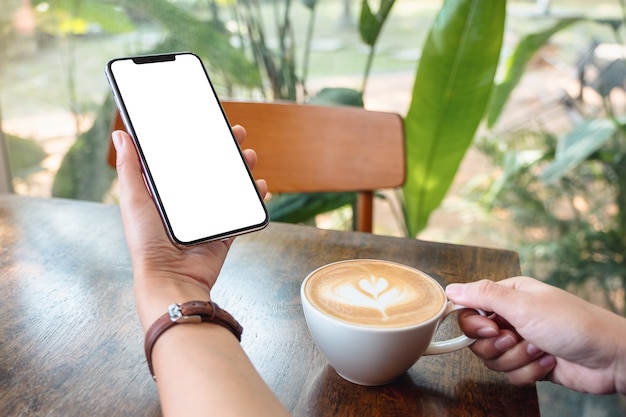 Modellbild einer Frau, die ein schwarzes Mobiltelefon mit leerem Desktop-Bildschirm hält und benutzt, während sie Kaffee im Café trinkt