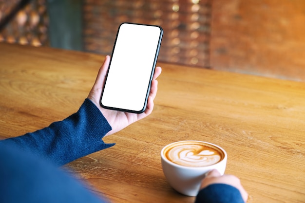 Modellbild einer Frau, die ein weißes Handy mit leerem Bildschirm hält, während sie Kaffee auf einem Holztisch im Café trinkt