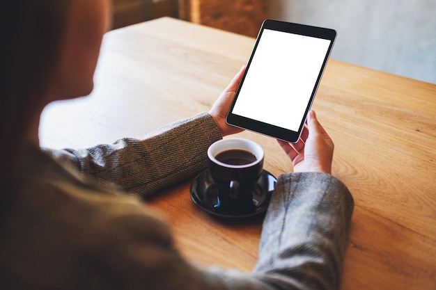 Modellbild einer Frau, die einen schwarzen Tablet-PC mit einem leeren weißen Bildschirm mit einer Kaffeetasse auf einem Holztisch hält