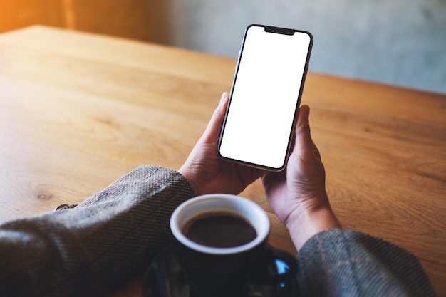 Modellbild einer Frau, die Handy mit leerem weißem Bildschirm mit Kaffeetasse auf Holztisch hält