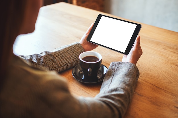 Modellbild einer Frau, die schwarzen Tablet-PC mit leerem weißem Bildschirm mit Kaffeetasse auf Holztisch hält