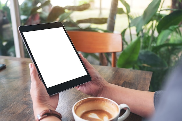 Modellbild einer Frau, die schwarzen Tablet-PC mit leerem weißem Bildschirm mit Kaffeetasse auf Holztisch hält