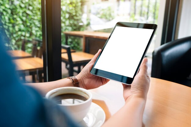 Modellbild von Händen, die schwarzen Tablet-PC mit leerem weißem Bildschirm mit Kaffeetasse auf Holztisch halten