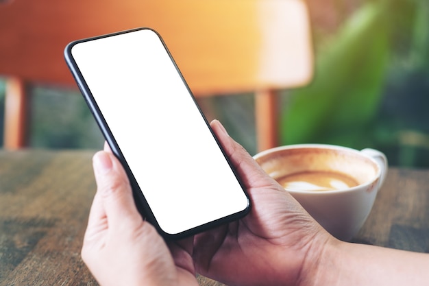 Modellbild von Händen, die schwarzes Handy mit leerem Bildschirm mit Kaffeetasse auf Holztisch im Café halten