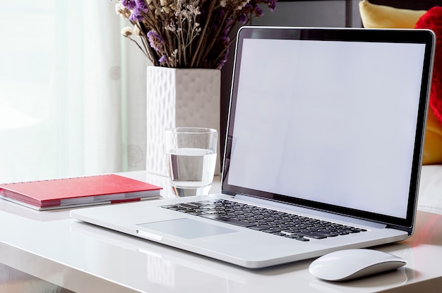 Modelllaptop mit leerem bildschirm, houseplant und tagebuch auf weißer spitzentabelle im wohnzimmer, leerer bildschirm für grafikdesign.