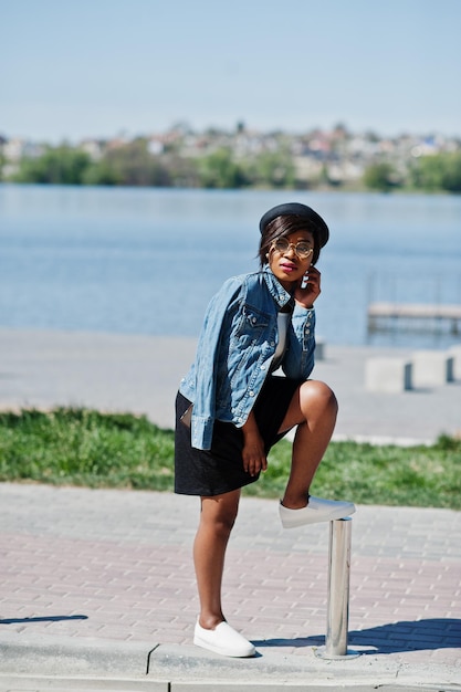 Foto modelo afro-americano elegante em óculos chapéu jaqueta jeans e saia preta posada ao ar livre