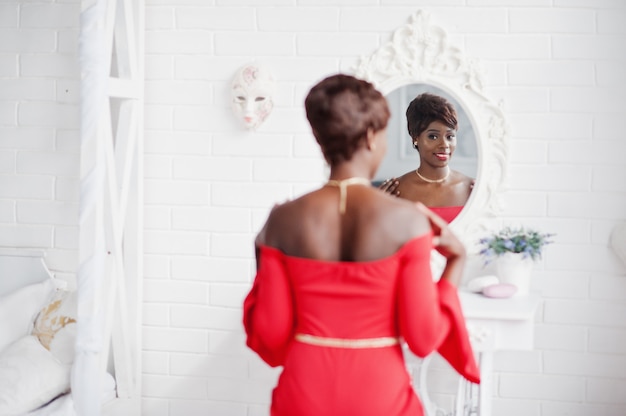 Modelo afroamericano de moda en vestido rojo de belleza, mujer sexy posando vestido de noche y mirando en el espejo.