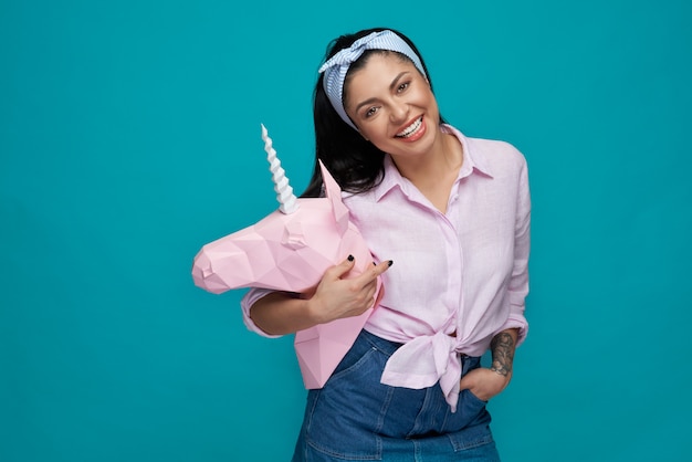 Modelo alegre posando, sosteniendo la cabeza de unicornio de papel rosa.