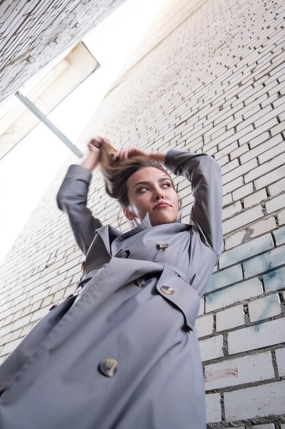 Modelo asiático atractivo en gabardina de pie cerca de la pared disparó desde el punto inferior