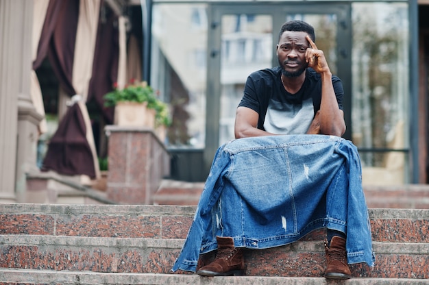 Modelo atractivo joven afroamericano serio. Chico guapo moderno en chaqueta de jeans sentado en la calle urbana de la ciudad.