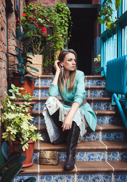 Foto modelo atractivo sentado en las escaleras interiores con ventanas panorámicas look primaveral en colores azul y beige