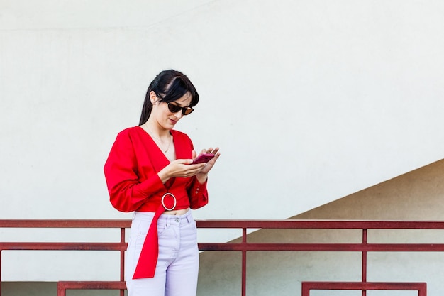 Modelo atraente de cabelo escuro em uma elegante roupa vermelha e branca olhando para o smartphone