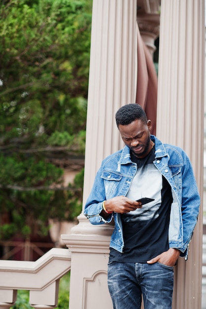 Modelo atraente jovem afro-americano sério. cara bonito moderno em jaqueta jeans em pé na rua da cidade urbana e olhando para o celular.