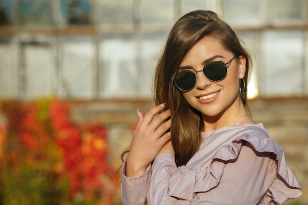 Modelo bronceado sonriente vistiendo blusa de moda y gafas de espejo, posando al aire libre con rayos de sol. Espacio para texto