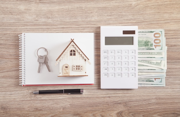 Modelo de casa de madera con teclas, calculadora, dinero, bolígrafo y bloc de notas.