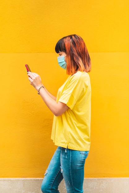 Modelo caucásico alternativo con cabello anaranjado caminando de lado mirando el teléfono móvil y con máscara